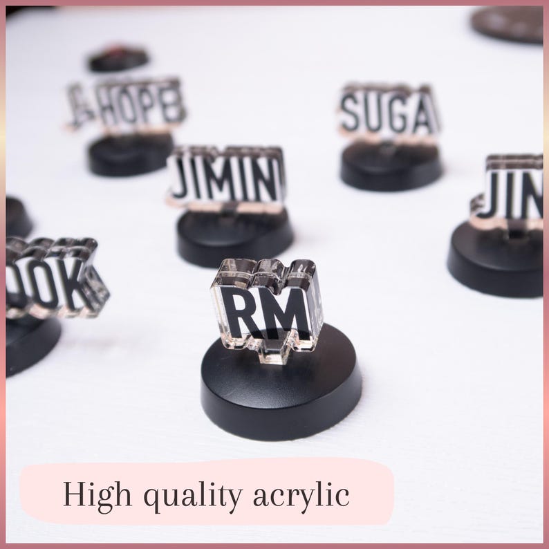May include: A collection of small, clear acrylic name plates featuring black text on white backgrounds, each mounted on a circular black base. The visible names include RM, J-HOPE, SUGA, JIMIN, JIN, and a partial view of COOK. The text High quality acrylic is present.