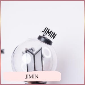 May include: A clear spherical light stick featuring a black base and a white BTS logo inside the globe. A small rectangular acrylic charm with the black text JIMIN is attached to the top of the black base, standing upright against the sphere.