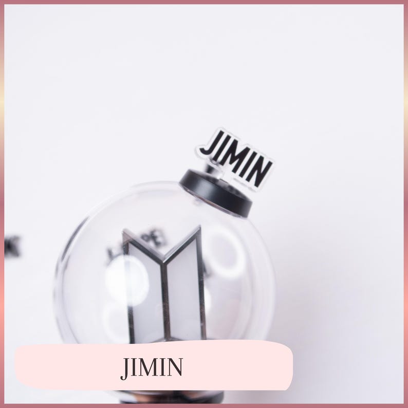 May include: A clear spherical light stick featuring a black base and a white BTS logo inside the globe. A small rectangular acrylic charm with the black text JIMIN is attached to the top of the black base, standing upright against the sphere.