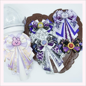 May include: A collection of decorative bows in shades of purple, white, and black, featuring character designs. The bows are arranged on a brown heart-shaped base. Some bows have gold accents and lace details.