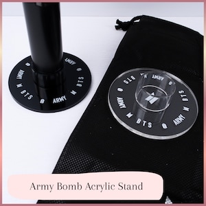 Könnte beinhalten: Schwarzer Acrylständer für eine Army Bomb, mit den Wörtern "BTS" und "ARMY" in weißer Schrift um die kreisförmige Basis. Ein klarer Acrylhalter befindet sich in der Mitte. Ein schwarzer Kordelzugbeutel ist ebenfalls enthalten.