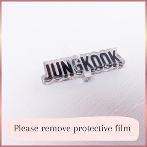 May include: A clear acrylic name tag featuring the black text JUNGKOOK in a bold, sans-serif font. The piece has a transparent, layered appearance with a partially peeled protective film on the surface. The design is rectangular and flat.