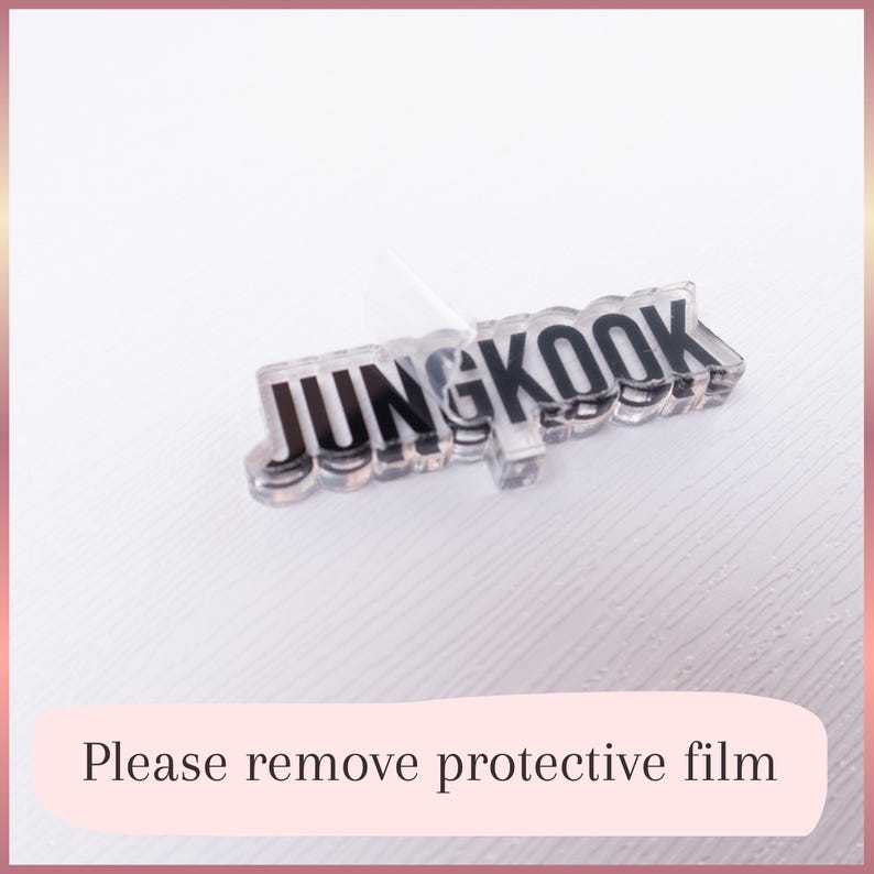 May include: A clear acrylic name tag featuring the black text JUNGKOOK in a bold, sans-serif font. The piece has a transparent, layered appearance with a partially peeled protective film on the surface. The design is rectangular and flat.