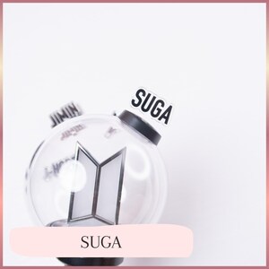 May include: A clear, spherical plastic light stick featuring a black base and a central silver BTS logo. A small, rectangular white tag with the black text SUGA is attached to the top. The word JIMIN is partially visible on a tag in the background.