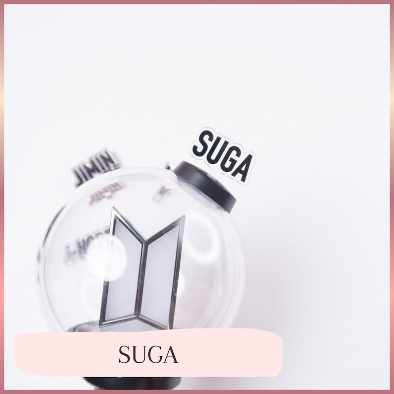 May include: A clear, spherical plastic light stick featuring a black base and a central silver BTS logo. A small, rectangular white tag with the black text SUGA is attached to the top. The word JIMIN is partially visible on a tag in the background.