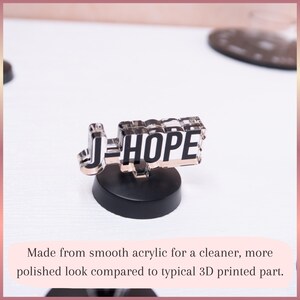 May include: A clear acrylic display piece featuring the black text J-HOPE in a bold, block font. The acrylic nameplate is mounted on a solid, circular black base. The design has a smooth, reflective surface and a transparent, layered appearance.