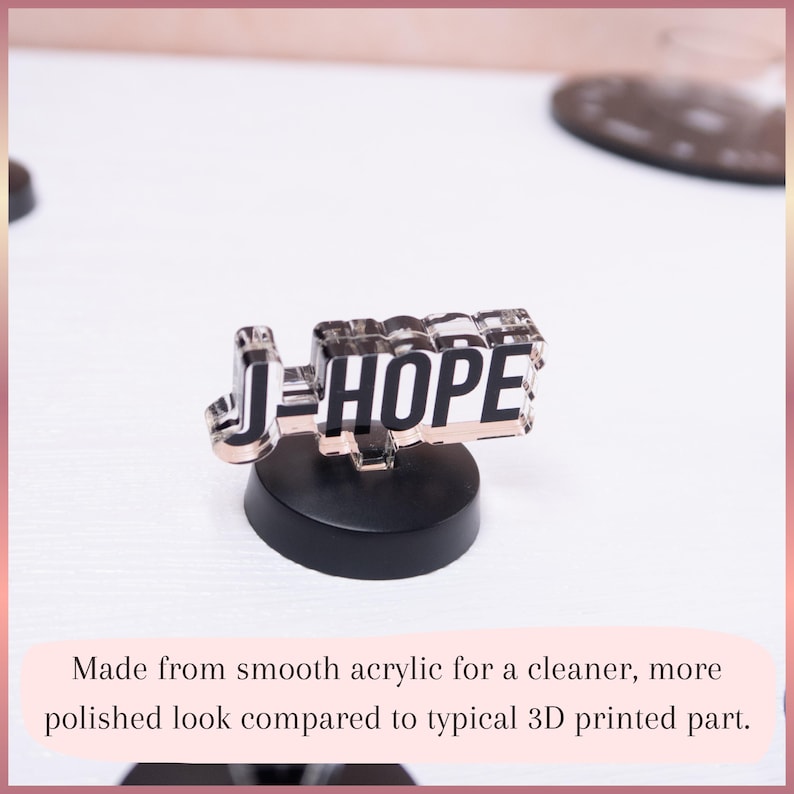 May include: A clear acrylic display piece featuring the black text J-HOPE in a bold, block font. The acrylic nameplate is mounted on a solid, circular black base. The design has a smooth, reflective surface and a transparent, layered appearance.
