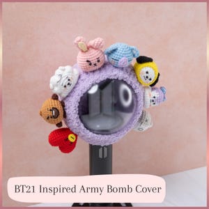 Puede incluir: Una funda de ganchillo lavanda y difusa para un bastón de luz circular, con ocho pequeñas cabezas de personajes coloridas y rellenas unidas alrededor del borde. El texto BT21 Inspired Army Bomb Cover aparece en la parte inferior.