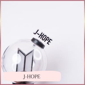 May include: A clear, spherical plastic light stick featuring a black base and a central geometric logo. A white rectangular tag with the black text J-HOPE is attached to the top of the black cap. The design is transparent and minimalist.