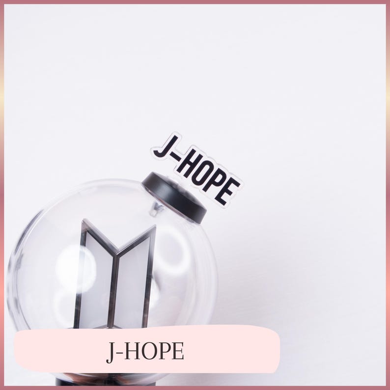 May include: A clear, spherical plastic light stick featuring a black base and a central geometric logo. A white rectangular tag with the black text J-HOPE is attached to the top of the black cap. The design is transparent and minimalist.