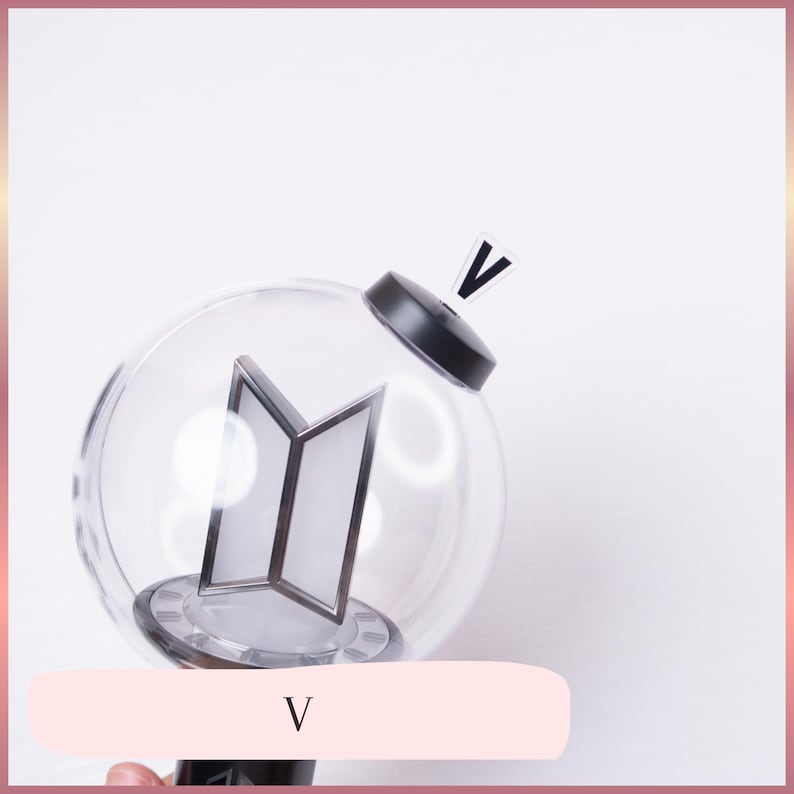 May include: A clear spherical light stick featuring a black base and a black top cap with a small V-shaped attachment. Inside the transparent globe, a silver-toned BTS logo is positioned vertically. The handle is black and cylindrical in shape.