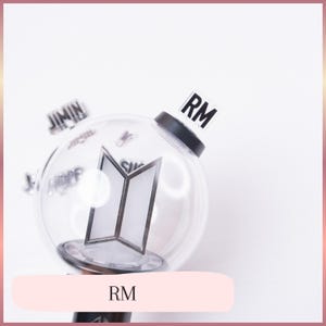 May include: A clear, spherical plastic light stick featuring a black base and a central white BTS logo emblem. A small, rectangular, transparent tab with the black text RM is attached to the top. Additional blurred name tags are visible in the background.