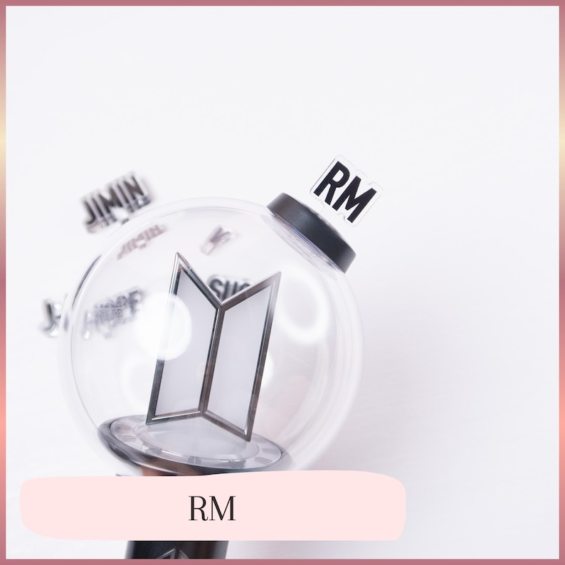 May include: A clear, spherical plastic light stick featuring a black base and a central white BTS logo emblem. A small, rectangular, transparent tab with the black text RM is attached to the top. Additional blurred name tags are visible in the background.