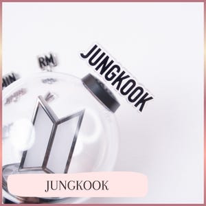 May include: A clear, spherical plastic light stick featuring a prominent BTS logo inside. A black topper with the white text JUNGKOOK is attached to the top. Additional small black charms with names like RM and JIMIN are visible inside the clear sphere.
