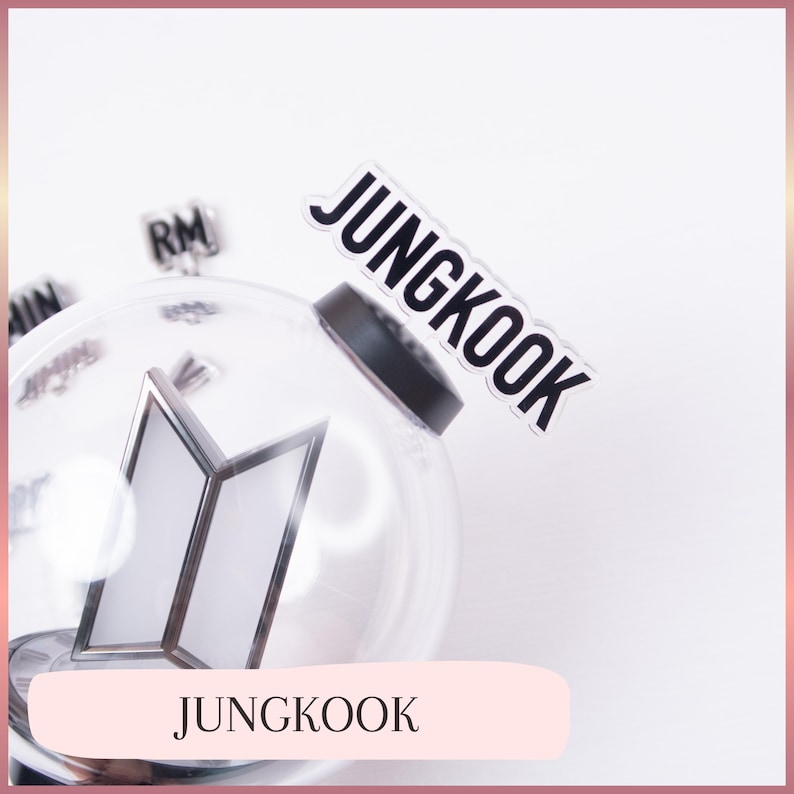 May include: A clear, spherical plastic light stick featuring a prominent BTS logo inside. A black topper with the white text JUNGKOOK is attached to the top. Additional small black charms with names like RM and JIMIN are visible inside the clear sphere.