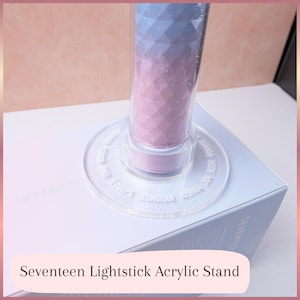 May include: A clear acrylic stand for a Seventeen lightstick. The stand has a circular base with engraved names and supports a lightstick with a purple and blue gradient. The stand is on a white box.