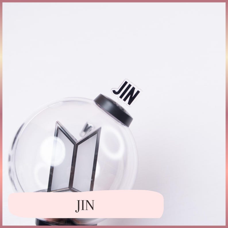 May include: A clear spherical light stick featuring a black base and a black BTS logo inside the globe. A small, rectangular, transparent sticker with the black text JIN is attached to the top of the black base. The design is simple and functional.