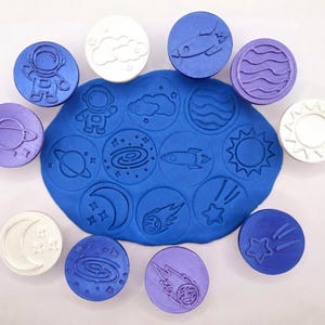 May include: A set of circular stamps featuring space-themed designs including an astronaut, rocket, planet, sun, moon, and stars, arranged around a central blue piece of modeling clay that displays the embossed impressions of these same shapes.
