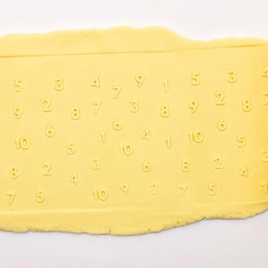 May include: A bright yellow cylindrical rolling pin with an embossed pattern of numbers from one to ten. The rolling pin is positioned next to a flat, light yellow sheet of dough that has been imprinted with the same numerical pattern.
