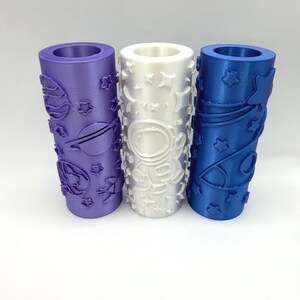 May include: Three cylindrical rollers in purple, white, and blue, each featuring embossed space-themed designs. The purple roller displays planets and stars, the white roller features an astronaut and stars, and the blue roller shows rockets and stars.