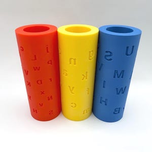 Op de afbeelding: Drie kleurrijke cilinders in rood, geel en blauw. Elke cilinder heeft een holle kern en is bedrukt met letters en cijfers. De rode cilinder heeft de letters V, W, D, K, X, H en de cijfers 2, 1. De gele cilinder heeft de letters S, K, C en de blauwe U, M, W, H en het cijfer 2.
