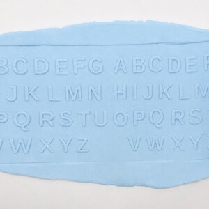 May include: A bright blue cylindrical rolling pin with embossed letters of the alphabet, shown pressing a repeating pattern of the letters A through Z into a flat, light blue piece of pliable material, creating a clear, indented impression.