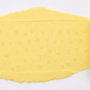 Op de afbeelding: Een felgele cilindrische deegroller met in reli&euml;f aangebrachte kleine letters van het alfabet. De deegroller ligt op een platte, lichtgele lap deeg en laat een duidelijk, ingedrukt patroon van diverse letters op het oppervlak achter.
