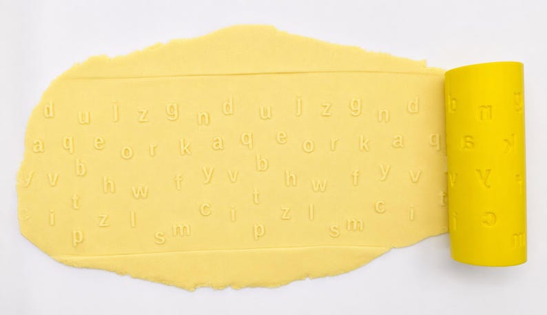 Op de afbeelding: Een felgele cilindrische deegroller met in reli&euml;f aangebrachte kleine letters van het alfabet. De deegroller ligt op een platte, lichtgele lap deeg en laat een duidelijk, ingedrukt patroon van diverse letters op het oppervlak achter.