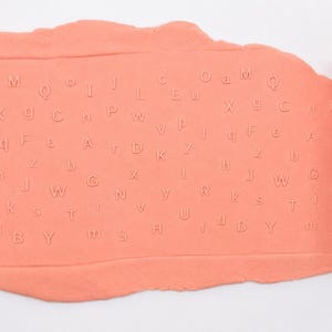 Op de afbeelding: Een cilindrische oranje deegroller met een reli&euml;fpatroon van het alfabet naast een platgeslagen vel perzikkleurige boetseerklei. De klei vertoont een duidelijke, ingedrukte afdruk van diverse hoofdletters en kleine letters.