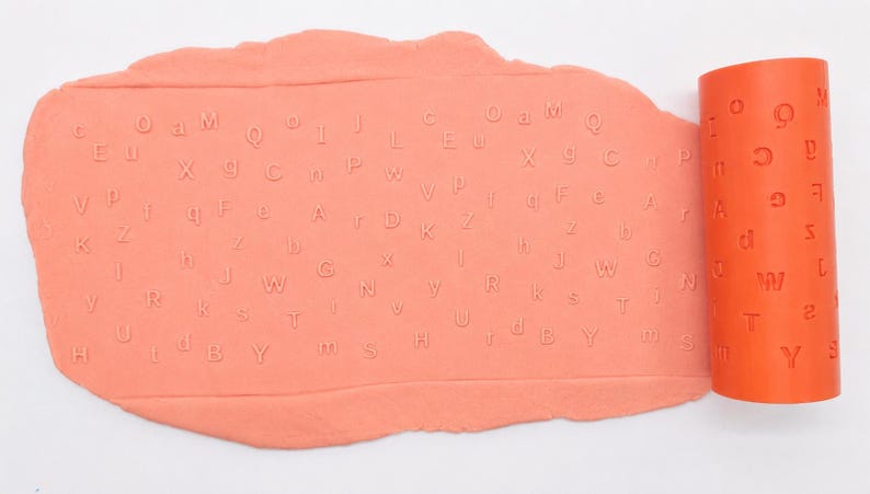 Op de afbeelding: Een cilindrische oranje deegroller met een reli&euml;fpatroon van het alfabet naast een platgeslagen vel perzikkleurige boetseerklei. De klei vertoont een duidelijke, ingedrukte afdruk van diverse hoofdletters en kleine letters.