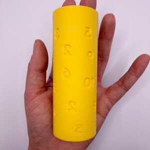 May include: A bright yellow cylindrical roller featuring embossed numerical characters scattered across its surface. The tool has a smooth, matte finish and is designed for creating impressions in soft materials, displaying various digits in relief.