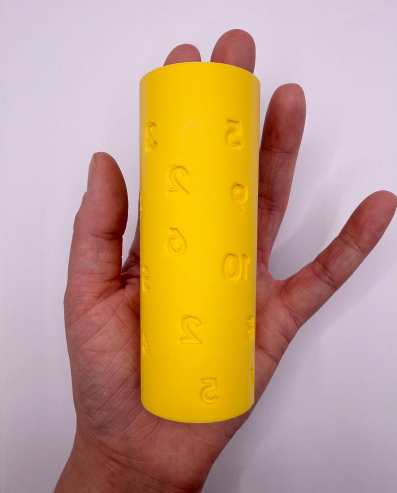 May include: A bright yellow cylindrical roller featuring embossed numerical characters scattered across its surface. The tool has a smooth, matte finish and is designed for creating impressions in soft materials, displaying various digits in relief.