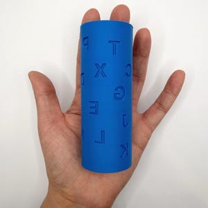 Op de afbeelding: Een felblauwe cilindrische roller met in reli&euml;f aangebrachte letters en cijfers op het oppervlak. De zichtbare tekens zijn onder andere P, T, X, C, D, G, E, 1, M, L en K, die in het materiaal zijn gedrukt. Het object heeft een gladde, matte afwerking.