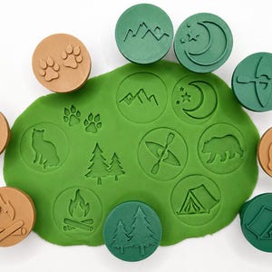 Puede incluir: Un conjunto de sellos redondos de plástico color canela y verde con iconos de naturaleza que incluyen un zorro, huellas, fogata, montañas, luna, kayak, oso y pinos, con sus impresiones marcadas en una base de masa verde.