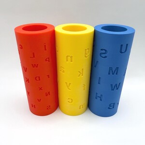 Op de afbeelding: Drie cilindrische rollers in rood, geel en blauw, elk voorzien van in reli&euml;f aangebrachte letters en cijfers op hun oppervlak. Deze holle buizen staan naast elkaar en tonen een verscheidenheid aan tekens, waaronder A, B, H, I, M, U en 2.