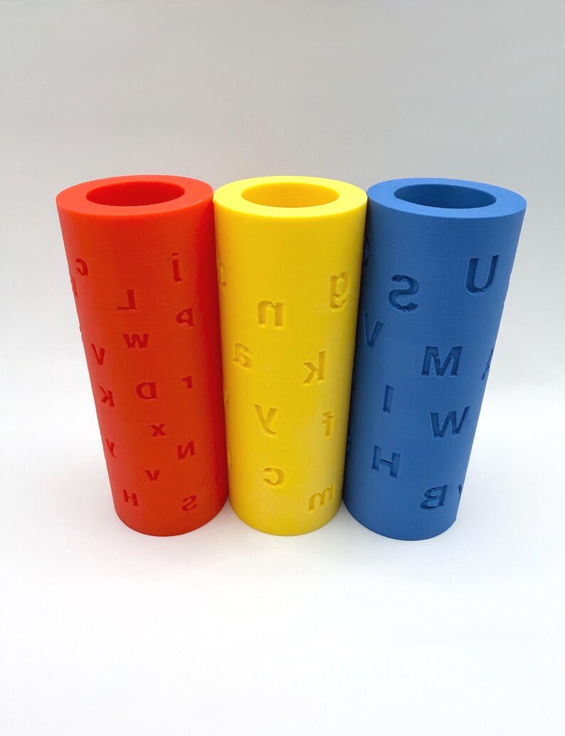 Op de afbeelding: Drie cilindrische rollers in rood, geel en blauw, elk voorzien van in reli&euml;f aangebrachte letters en cijfers op hun oppervlak. Deze holle buizen staan naast elkaar en tonen een verscheidenheid aan tekens, waaronder A, B, H, I, M, U en 2.