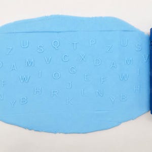 Op de afbeelding: Een lichtblauwe cilindrische deegroller met verdiepte letters van het alfabet, geplaatst naast een platgeslagen vel lichtblauw deeg dat de reli&euml;fafdrukken van de letters P, Z, U, S, Q, T, X, D, A, M, V, C, E, F, W, G, J, H, R, L, N, B en K toont.