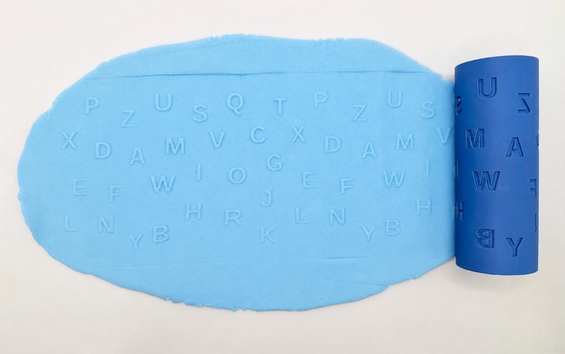 Op de afbeelding: Een lichtblauwe cilindrische deegroller met verdiepte letters van het alfabet, geplaatst naast een platgeslagen vel lichtblauw deeg dat de reli&euml;fafdrukken van de letters P, Z, U, S, Q, T, X, D, A, M, V, C, E, F, W, G, J, H, R, L, N, B en K toont.