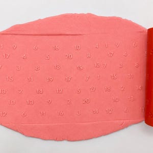 May include: A flat, oval-shaped piece of pink modeling clay featuring an embossed pattern of scattered numbers from 1 to 20. Beside the clay is an orange cylindrical rolling pin with the same numerical pattern engraved on its surface.