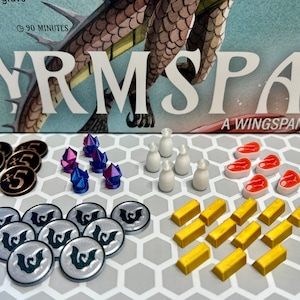 Könnte beinhalten: Eine Sammlung von Spielsteinen für das Brettspiel "Nyrmspan, A Wingspan Game". Die Spielsteine umfassen Goldbarren, silberne Spielsteine mit einem Flügel-Symbol, rote Fleisch-Spielsteine, weiße Milch-Spielsteine und lila und blaue Kristalle.