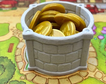 May include: A white, stone-like well with a brick pattern, filled with gold coins. The well is sitting on a green and brown game board.