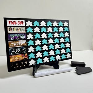 Puede incluir: Un tablero rectangular negro con siete filas horizontales de títulos de juegos que incluyen Final Girl, Root, Boooop, Terraforming Mars, Arcs, Slay the Spire y Dune Imperium, con filas de meeples de madera blancos y turquesas, un soporte y un marcador blanco.