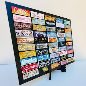 以下が含まれることがあります： 「Catan」、「Candy Land」、「Gloomhaven」などのボードゲームのタイトルが並んだ額入りのディスプレイ。タイトルはグリッド状に配置され、黒いフレームとスタンドが付いています。画像は角度がついており、さまざまなゲームが表示されています。