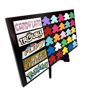 以下が含まれることがあります： Candy Land、Trouble、Flip 7、Everdell、Pokémonというタイトルの5つの長方形のプレートが付いた黒いディスプレイボード。ボードには、さまざまな単色の小さな人型のゲームピースが多数配置されています。