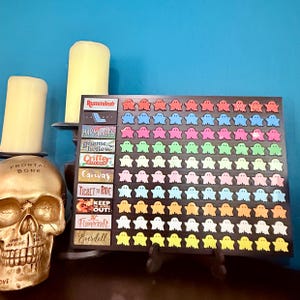 Puede incluir: Un tablero negro con piezas de juego en forma de fantasma de colores. El tablero tiene etiquetas de texto para varios juegos, incluyendo "Rummikub", "Harmonies" y "Ticket to Ride". Una calavera dorada y velas están en el fondo.