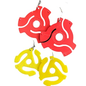 May include: Red and yellow vinyl record adapter earrings. The earrings are made of plastic and feature a circular design with a hole in the center. The yellow earrings have the text "ADAPTOR 45 R.P.M." printed on them. The earrings hang from silver-colored hooks.