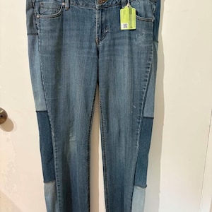 May include: Blue denim jeans featuring a patchwork design with varying shades of blue fabric panels. The jeans have a standard waistband with a button closure, front pockets, and a light green rectangular tag attached to the belt loop.