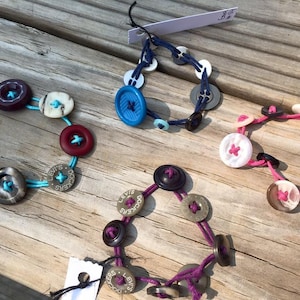 May include: Four bracelets made with various buttons and string. The bracelets are in different colors, including blue, pink, red, and purple. The buttons are made of different materials, including plastic, metal, and wood.