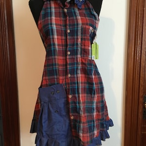 May include: A halter-style apron made from a repurposed plaid shirt. The apron features a red, blue, and green plaid pattern, with a denim pocket and ruffled hem. A blue flower adorns the neckline. Buttons run down the front.