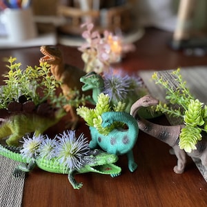 Puede incluir: Una colección de dinosaurios de juguete en varios colores, incluyendo verde, marrón y turquesa, adornados con pequeñas plantas verdes y flores moradas. Los dinosaurios están dispuestos sobre una superficie de madera.